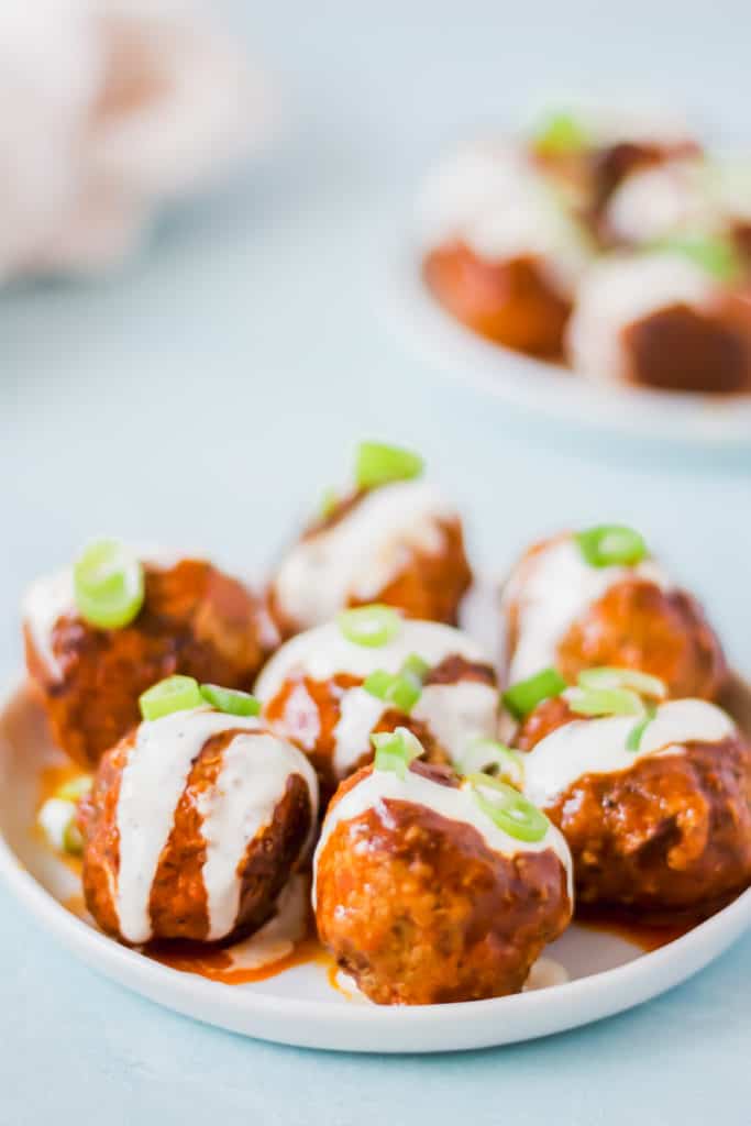 orange colored meatballs on a white plate with white ranch dressing and green onions on top with a blue background and white napkin