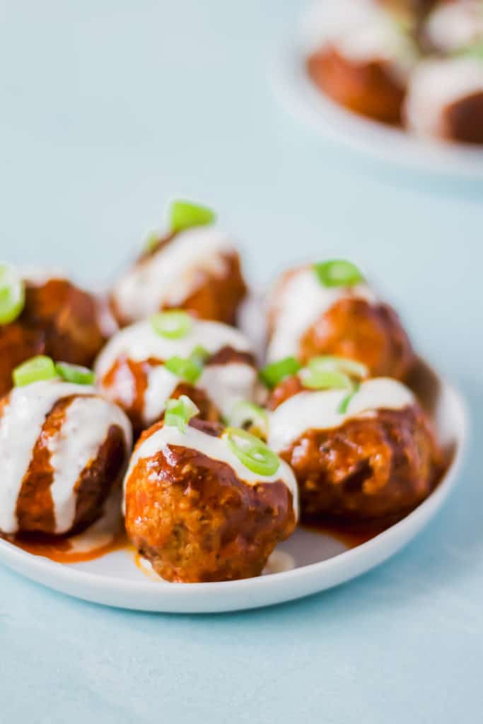 seven meatballs arranged on a white plate drizzled with white ranch dressing and green onion on a light blue background