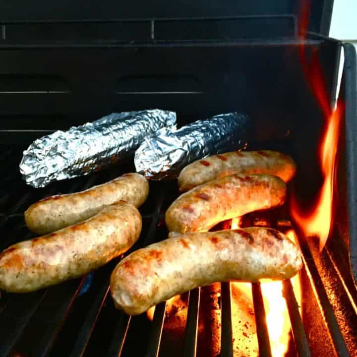 brats and corn wrapped in aluminum foil on a hot grill