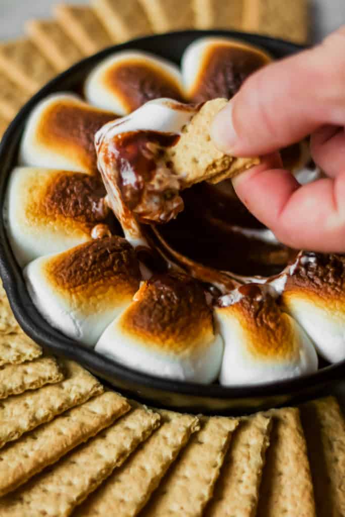 graham cracker being dipped into marshmallows and chocolate in a cast iron skillet surrounded by a circle of graham crackers