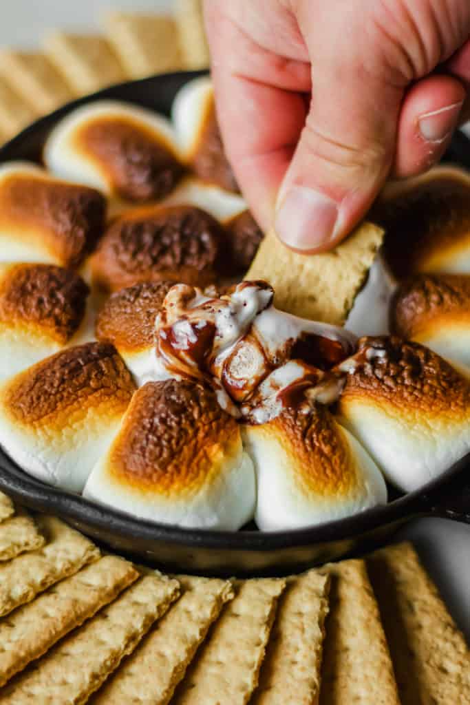 a person holding a graham cracker dipping it into a cast iron filled with browned marshmallows