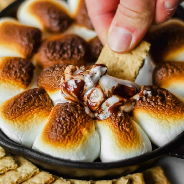 a person holding a graham cracker dipping it into a cast iron filled with browned marshmallows