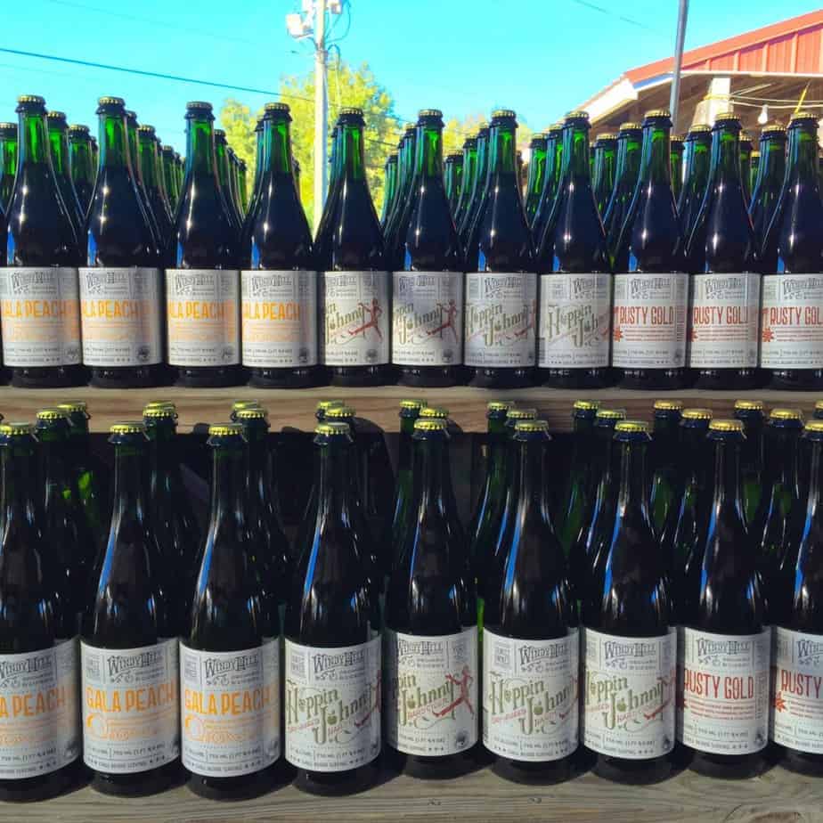 Rows of bottled craft beers from Windy Hill Orchard line wooden shelves, with green glass bottles labeled Galad Peach, Heppin Johnny, and Rusty Gold. Sunlight and blue sky are visible in the background.