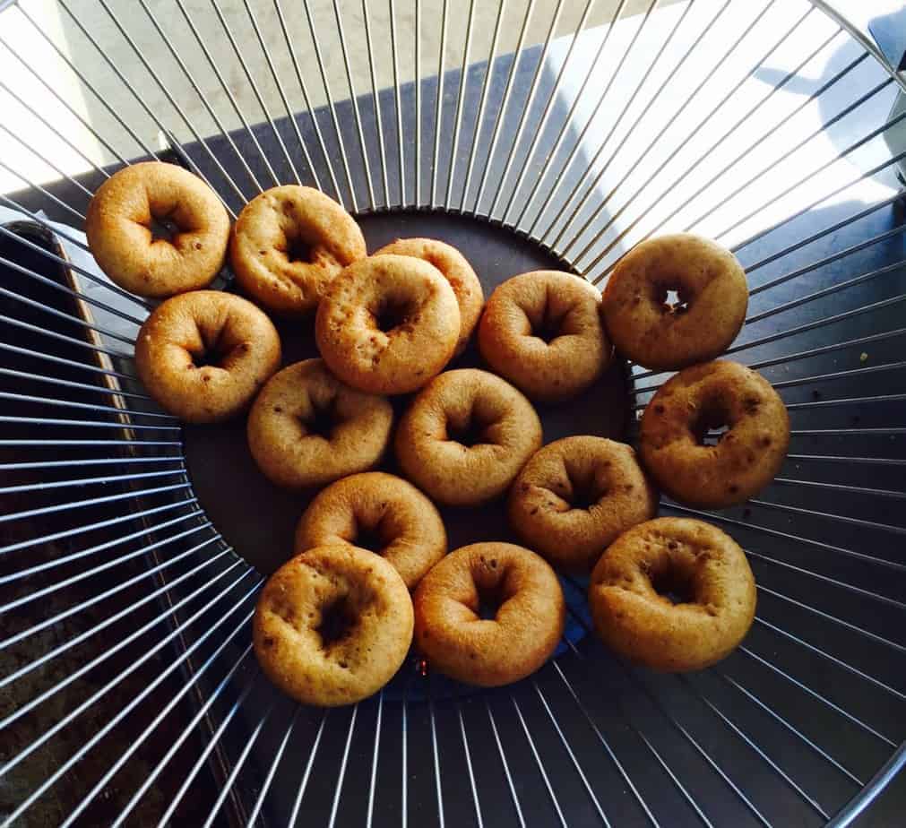 A group of small, round bagels or doughnuts from Windy Hill Orchard are arranged on a circular wire cooling rack, with sunlight casting delicate shadows through the rack.