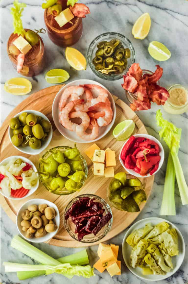 a diy bloody mary bar on a marble and wooden board with toppings and bloody mary bar ingredients