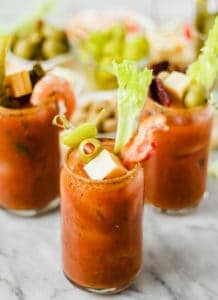 Three Bloody Mary cocktails garnished with celery, cheese cubes, shrimp, olives, pickles, and cherry tomatoes sit on a marble surface—perfect inspiration for your next build your own Bloody Mary bar.