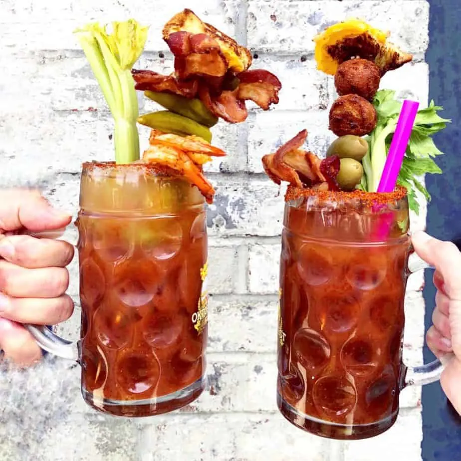 two large bloody marys with lots of toppings in front of a white wall