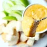 A fork holds a piece of bread dipped in beer cheese fondue, with sliced green apples, more bread cubes, and a bowl of cheesy dip in the background.