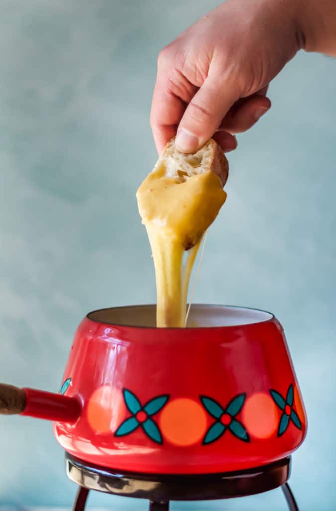 dipping bread into cheddar cheese fondue that is being served in a red orange and blue fondue pot