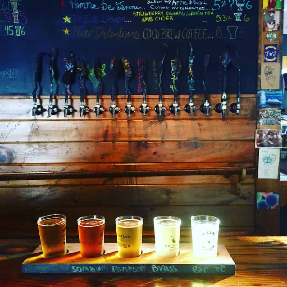 A wooden bar with four glasses of beer on a paddle, each a different color. Behind them are beer taps mounted on a wall decorated with chalkboard menus and colorful stickers—perfect for a honeymoon in Asheville.