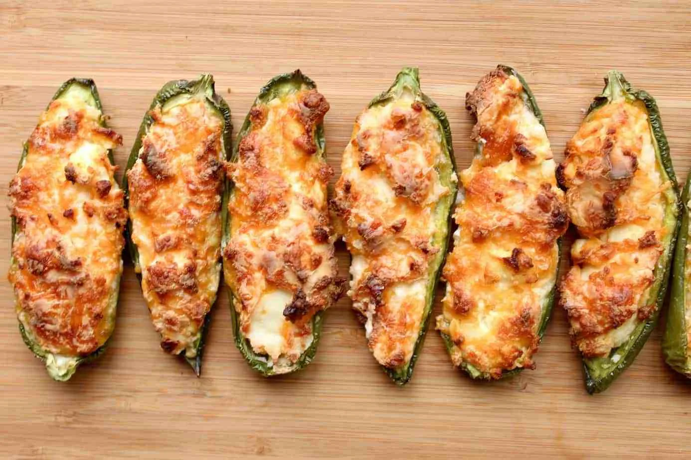 a line of baked jalapeno poppers with browned cheese on top on a wooden cutting board a line of baked jalapeno poppers with browned cheese on top on a wooden cutting board
