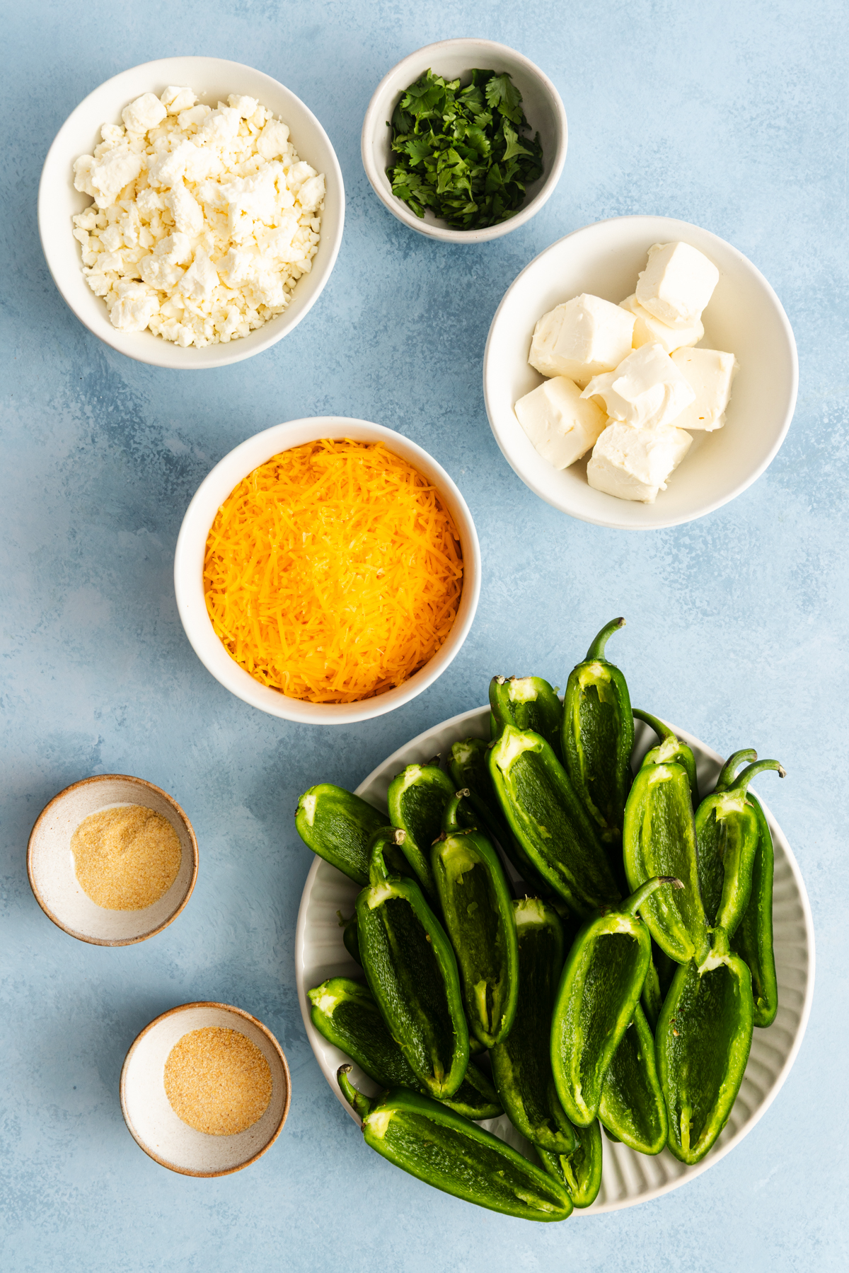 Ingredients for jalapeño poppers, including halved jalapeños, shredded cheddar, feta, cream cheese, chopped parsley, and spices in small bowls.
