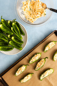 Jalapeño halves being stuffed with a cheese mixture on a parchment-lined baking sheet to prepare jalapeño poppers, with a bowl of filling and extra halved peppers nearby.