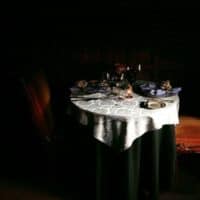 A small round table set for two with elegant plates, glasses, and folded napkins sits in dim lighting—an intimate scene reminiscent of Charlotte's romantic restaurants. A white lace tablecloth and ornate chairs complete the inviting setting.