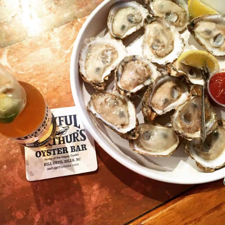 A plate of fresh oysters on the half shell with lemon wedges and cocktail sauce sits beside a bottle of Corona beer atop an Awful Arthur’s Oyster Bar coaster—a classic taste of Outer Banks travel.