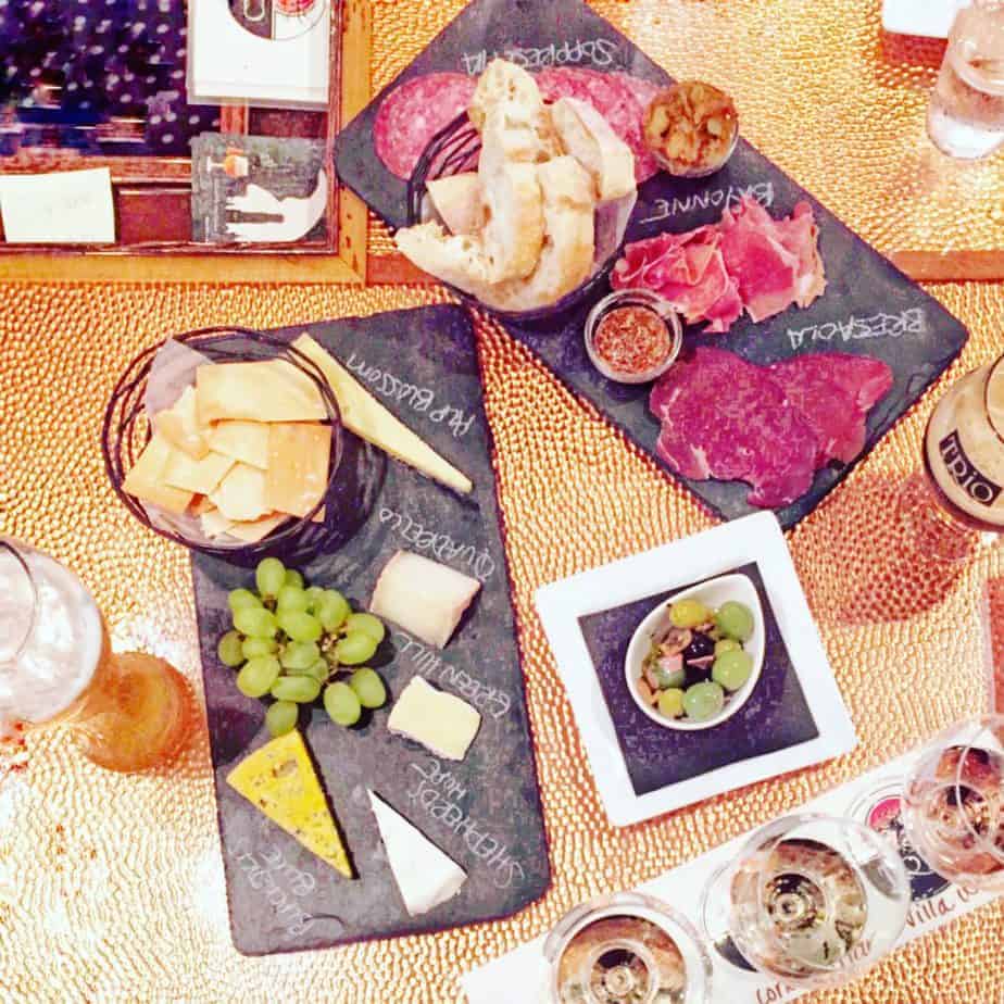 Charcuterie boards with assorted cheeses, grapes, crackers, sliced cured meats, bread, and bowls of olives and condiments on a gold textured table—perfect for savoring during your Outer Banks travel experience. Glasses and drinks surround the boards.