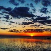 A vibrant sunset over a calm body of water at one of the beaches near Charlotte, NC, with dark clouds scattered across a colorful sky and the sun casting orange and yellow reflections on the rippling surface.