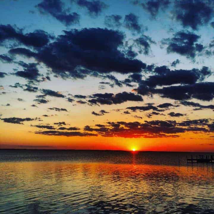 A vibrant sunset over a calm body of water at one of the beaches near Charlotte, NC, with dark clouds scattered across a colorful sky and the sun casting orange and yellow reflections on the rippling surface.