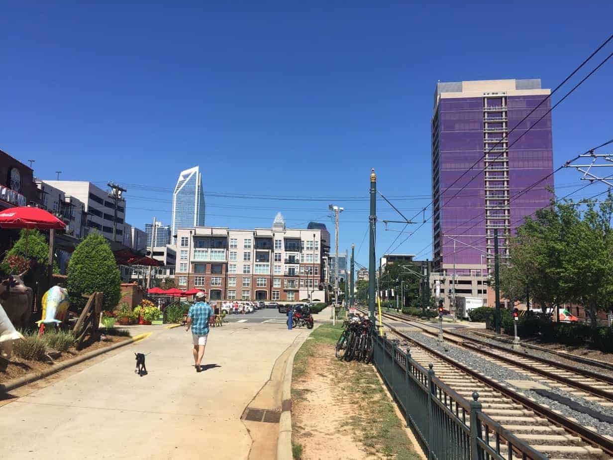 A man walks a dog beside train tracks in a city with modern buildings and a purple high-rise under a clear blue sky. Bicycles are parked by the fence and people sit at an outdoor café on the left.