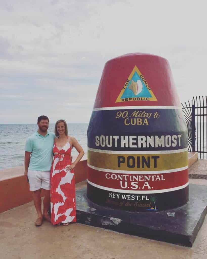 A couple stands by the large, colorful Southernmost Point Buoy in Key West, Florida, making a memorable stop on their Florida road trip. The ocean and sky are in the background, and the buoy reads 90 Miles to Cuba, Southernmost Point, Continental U.S.A..
