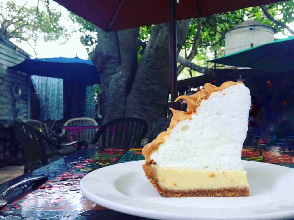 A slice of pie with a thick, fluffy meringue topping sits on a white plate outdoors, surrounded by patio furniture and shaded by large umbrellas and trees—evoking the charming ambiance often found at Key West restaurants.