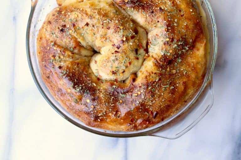 A golden-brown, spiral-shaped pepperoni bread topped with herbs and spices sits in a round glass baking dish on a white marble surface.