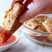 A hand dips a slice of pepperoni bread, filled with gooey cheese and spicy pepperoni, into a bowl of marinara sauce. In the background, a round baking dish holds more delicious stuffed bread slices.