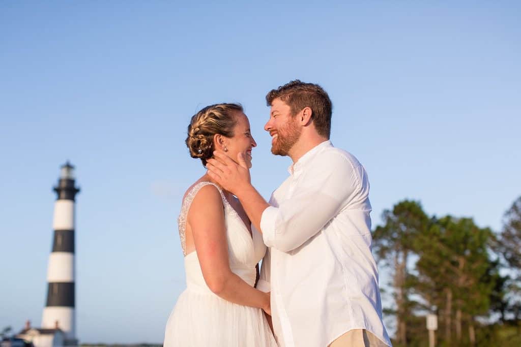 A couple in white clothing stands smiling and gazing at each other outdoors, with a lighthouse and trees in the background under a clear blue sky—capturing the magic of an Outer Banks wedding.
