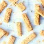 Golden-brown cheese straws are scattered on a white surface, with crumbs around them. Each cheese straw features a ridged texture and appears delightfully crispy and perfectly baked.
