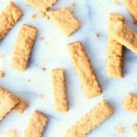 Golden-brown cheese straws are scattered on a white surface, with crumbs around them. Each cheese straw features a ridged texture and appears delightfully crispy and perfectly baked.