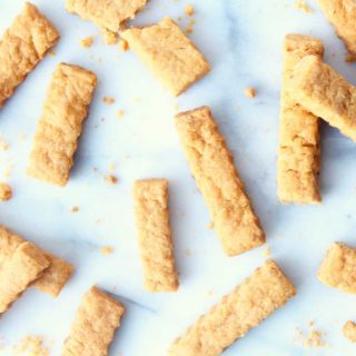 Golden-brown cheese straws are scattered on a white surface, with crumbs around them. Each cheese straw features a ridged texture and appears delightfully crispy and perfectly baked.