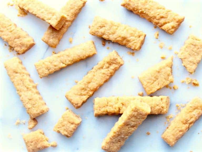 Rectangular pieces of golden, crumbly shortbread cookies are scattered on a white surface, with a few crumbs around them, resembling classic cheese straws in their shape and appeal.