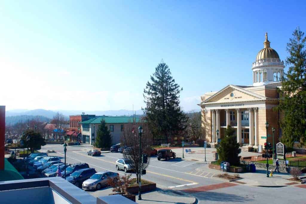 A small town street with parked cars, historic buildings, and a domed courthouse under a clear blue sky, with trees, distant hills, and charming Hendersonville restaurants lining the sidewalks.