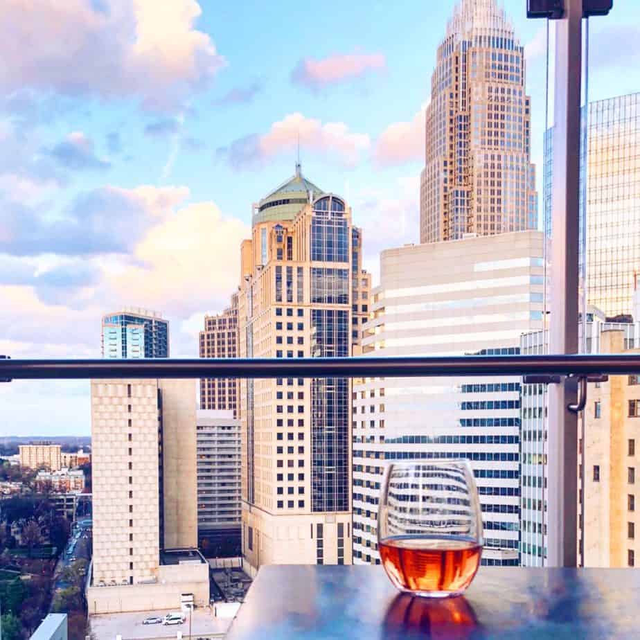 A glass of rosé wine sits on a table on a balcony overlooking a city skyline with tall buildings at sunset—a perfect way to unwind and enjoy one of the top things to do in Charlotte under a partly cloudy sky.