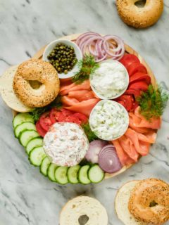 lox, cream cheese, tomatoes, cucumbers, onions, fresh dill, capers and bagels arranged on a wooden board with a marble background
