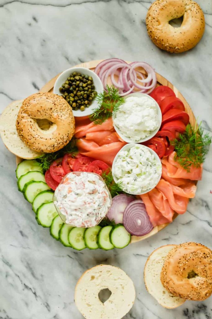 lox, cream cheese, tomatoes, cucumbers, onions, fresh dill, capers and bagels arranged on a wooden board with a marble background