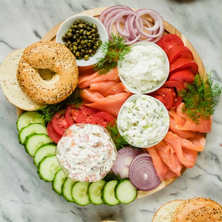 lox, cream cheese, tomatoes, cucumbers, onions, fresh dill, capers and bagels arranged on a wooden board with a marble background