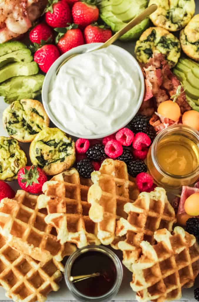 brunch board made with waffles, maple syrup, honey, egg muffins, raspberries and blackberries, greek yogurt, bacon and avocado