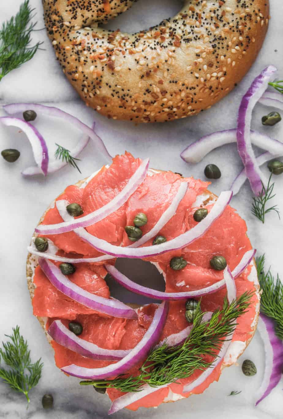 two halves of an everything bagel topped with lox, red onion and fresh dill on a marble background