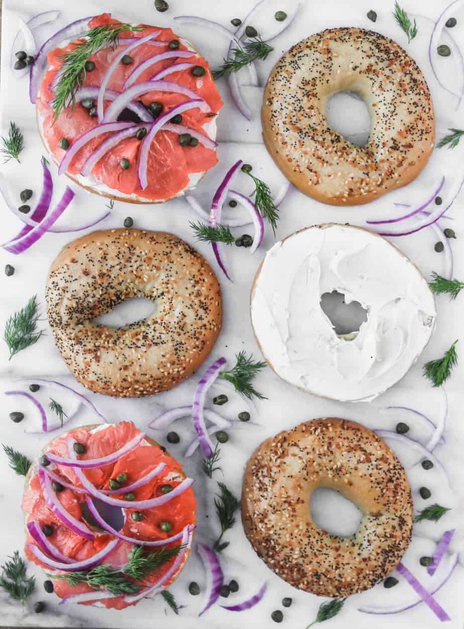 six bagel halves, two topped with pink smoked salmon and one topped with cream cheese on a marble background with red onions and herbs