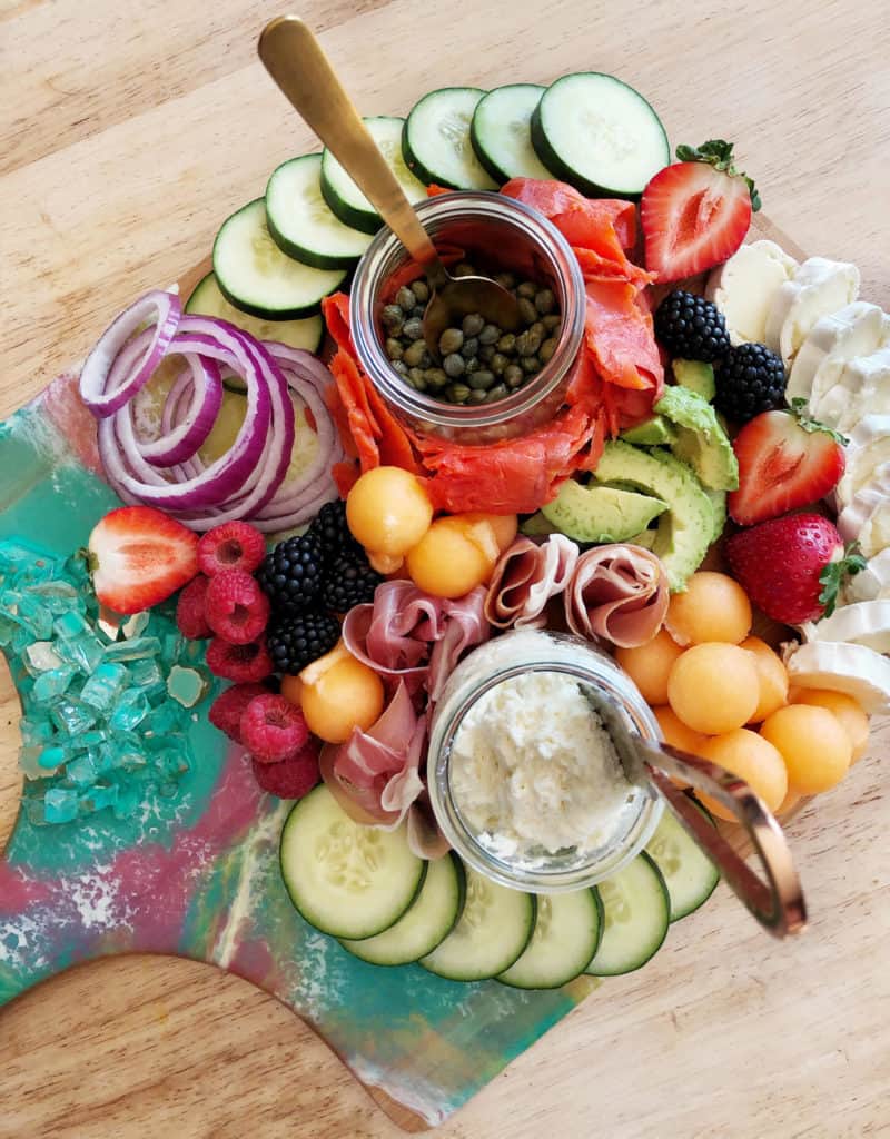 bagels and lox brunch board with cucumbers, red onion, melon, strawberries, avocado, brie cheese, raspberries and red onino on a pink and teal board with a wood background