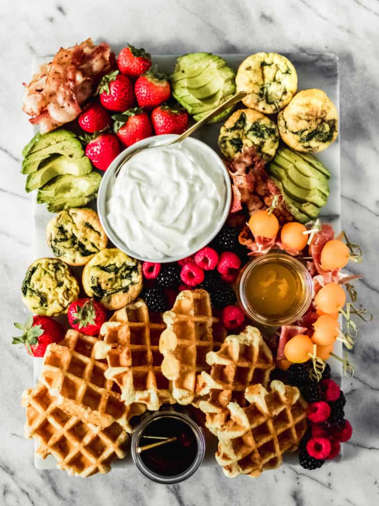 waffle themed brunch board with syrup, fresh fruit, avocado and eggs on a marble background