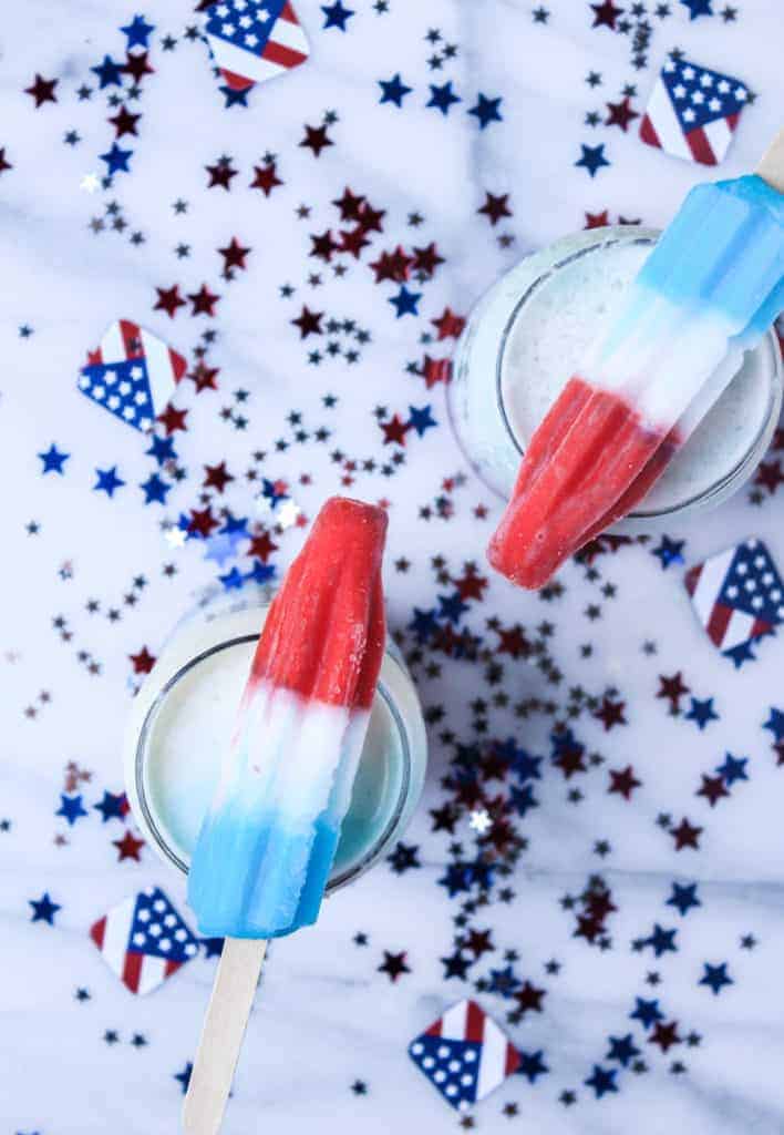 two cups filled with a 4th of july cocktail with red, white and blue popsicles on top on a marble background with red, white and blue confetti