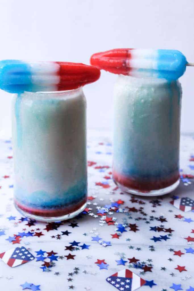 two glasses filled with a red white and blue 4th of july drink topped with popsicles on top of a white board with confetti