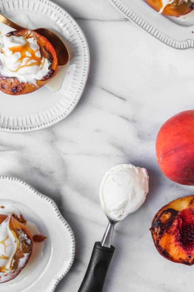 grilled peach halves made into sundaes with melted vanilla ice cream on white plates with an ice cream scoop, ice cream and whole peaches