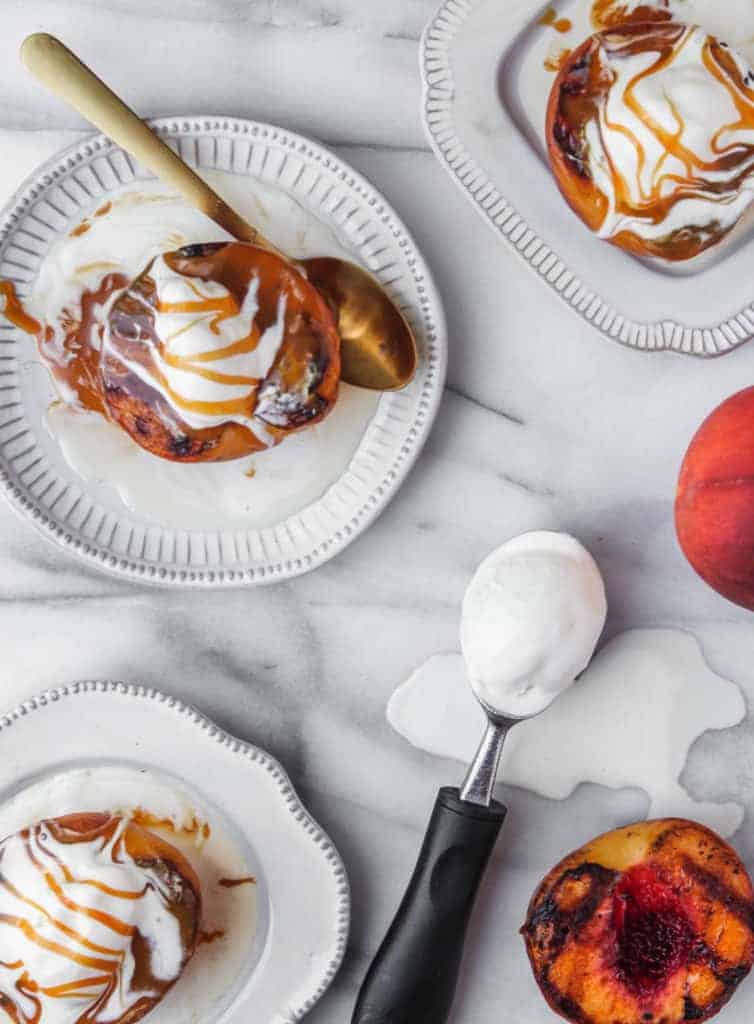 grilled peaches with ice cream and caramel sauce on white plates with a gold spoon. metal ice cream scoop with vanilla ice cream melting on a marble background.