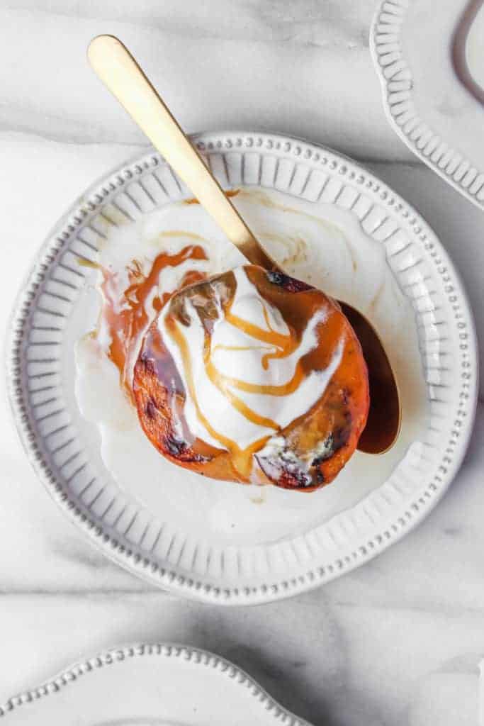 single grilled peach with vanilla ice cream and caramel sauce on a white plate with a gold spoon