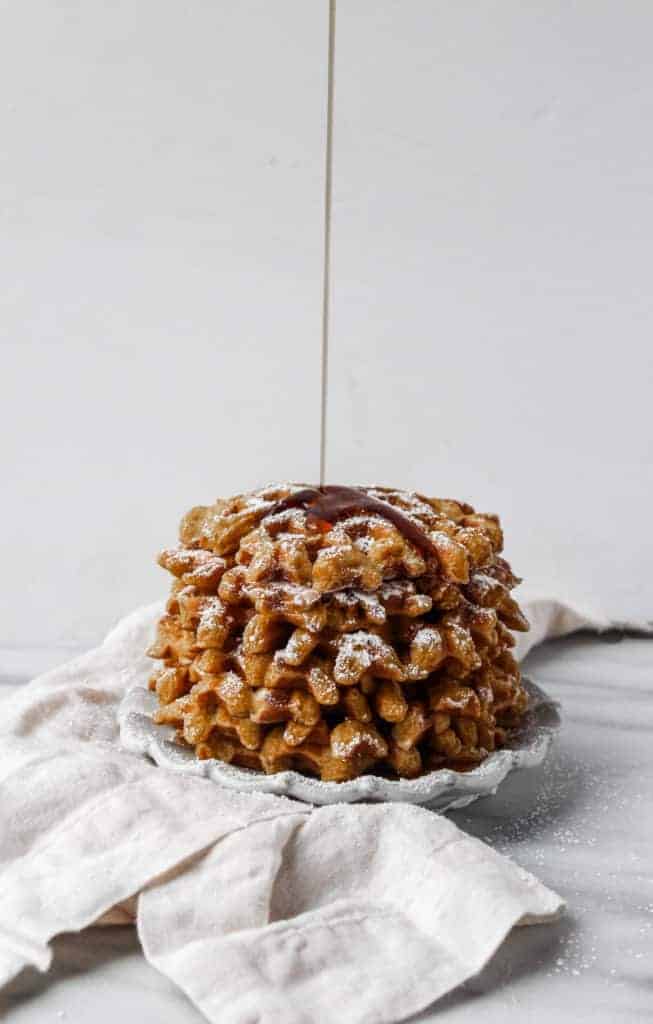 pouring syrup on a stack of pumpkin waffles that are stacked on a white plate next to a white napkin