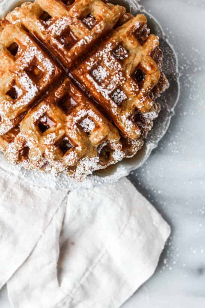 stack of pumpkin waffles on a white plate next to a white napkin topped with powdered sugar and syrup