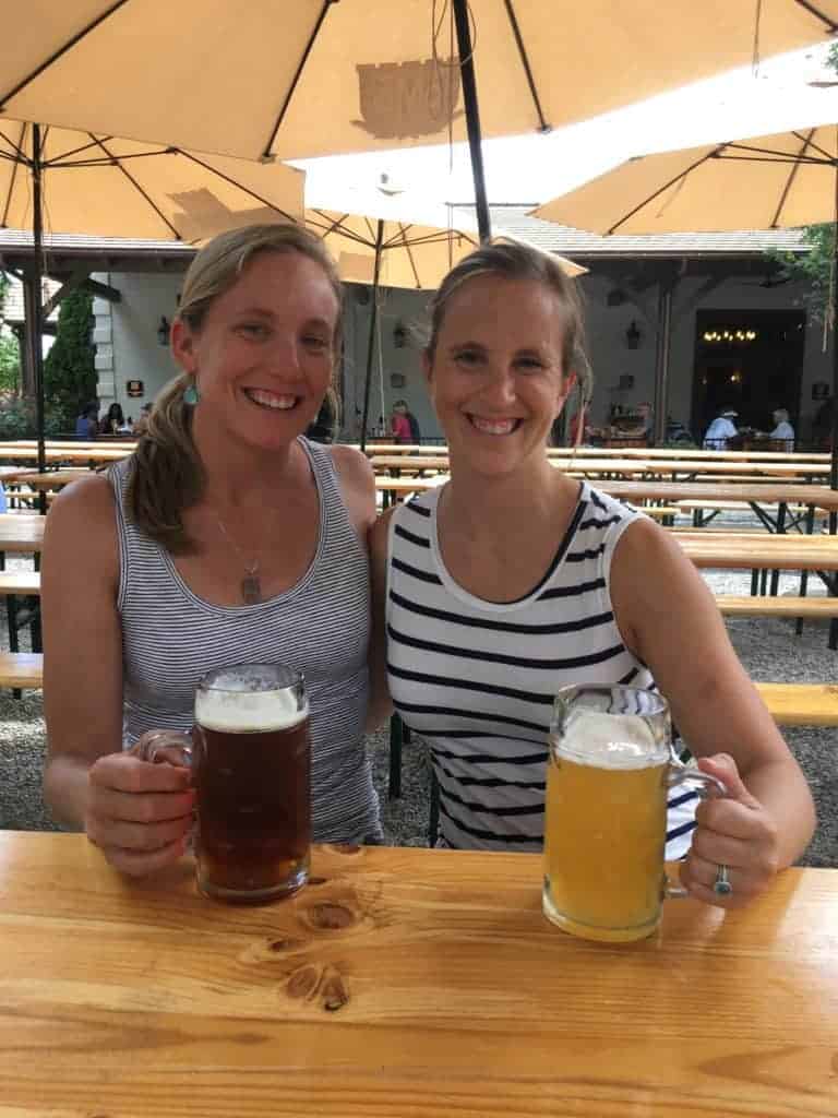 Two women sitting at an outdoor beer garden, smiling and holding large mugs of beer; one has a dark beer, the other a light beer. They sit at a wooden table under umbrellas on a sunny day.